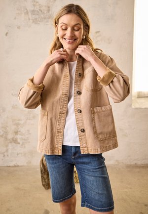 Smilende kvinde justerer knapperne på en beige jakke over en hvid skjorte, iført blå denimshorts, stående i et rum med neutrale farver og en tekstureret væg.