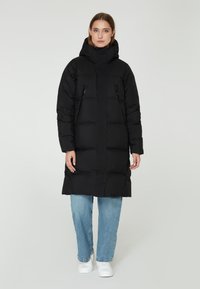 Parka negra acolchada con cuello alto y capucha, que cuenta con dos bolsillos laterales con cremallera, llevada sobre unos jeans azul claros y zapatillas blancas.