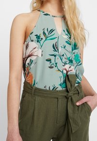 Top de halter con estampado floral en verde claro y un corte en forma de llave; combinado con pantalones de talle alto en verde oliva con detalle de cinchado en la cintura.