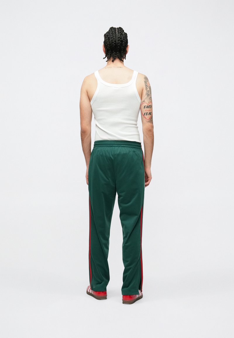 Pantalon de sport vert avec des bandes rouges sur les côtés, taille élastique et texture lisse, porté avec un débardeur blanc et des chaussures rouges.