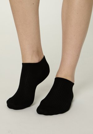 Füße mit kurzen schwarzen, gerippten Knöchelsocken vor hellem Hintergrund, ein Fuß leicht auf die Zehen gehoben.