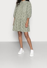 Ljust grönt kläddress med knäppning framtill, korta puffärmar och spetsliknande utskärningar. I kombination med vita sneakers på en neutral bakgrund.