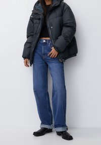 Puffet sort jakke med hætte, parret med højtaljede blå jeans med frynset kant, og sorte ankelstøvler på en ensfarvet baggrund.