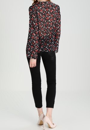 Donna che indossa una camicetta nera trasparente con motivo di labbra rosse e stelle bianche, pantaloni neri aderenti e scarpe beige con tacco alto, vista di schiena.