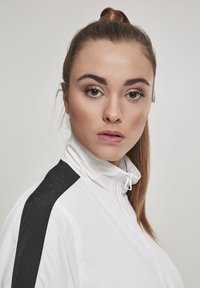 Bijela jakna na zip s visokim ovratnikom, s crnim detaljima na ramenima. Glatka tekstura, minimalistički dizajn i bez vidljivih uzoraka.