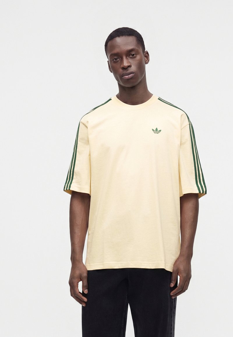 Νέος άνδρας που φοράει ένα κρεμ T-shirt Adidas με σκούρες πράσινες ρίγες στα μανίκια και ένα μικρό λογότυπο τριφύλλι στο στήθος, στέκεται μπροστά σε ένα απλό λευκό φόντο.