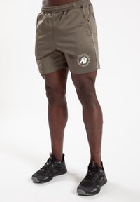 Olijfgroene sportshorts met elastische tailleband, voorzien van witte zijstrepen en een geprinte logo. Combinatie met zwarte hardloopschoenen.