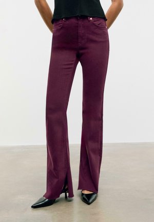 Jeans in denim bordeaux con gambe larghe e spacchi laterali all'orlo, abbinati a décolleté nere a punta. Texture liscia e design sartoriale.