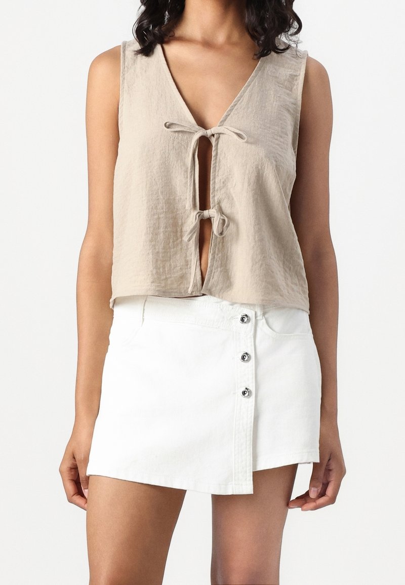 Haut court beige sans manches avec deux liens à l'avant, associé à une mini-jupe blanche boutonnée, porté par une personne debout.