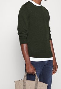 Mörkgrön stickad tröja med långa ärmar, rund halskrage och uppvikta handleder, lager på lager över en vit skjorta. En ljusgrå toteväska hålls.