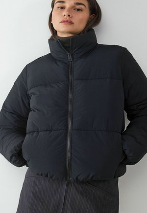 Schwarze Daunenjacke mit hohem Kragen, gestepptem Design und Reißverschluss vorne. Lange Ärmel und weiche, glatte Textur. Über einem dunkel gestreiften Rock getragen.