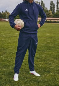 Sweat à capuche bleu marine et survêtement avec logo Adidas blanc ; le joueur tient un ballon de football usé, porte des crampons blancs et roses, debout sur l'herbe.