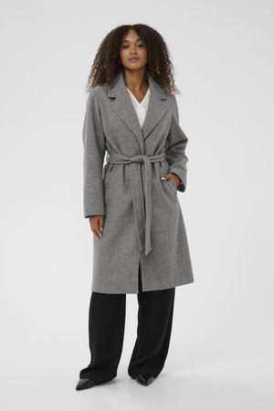 Grå ullblandad kappa med snedkrage, bältad midja, framknäppning med knappar och sidofickor, bärs över svarta vida byxor.