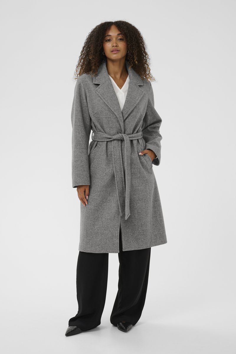 Sivý kabát z vlnenej zmesi s klopovým golierom, opaskom v páse, predným gombíkovým zapínaním a bočnými vreckami, nosený cez čierne nohavice s širokými nohavicami.
