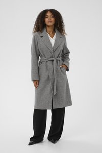 Halljas hall villasegu taks, millel on krae, vöökoht, ees nupuga sulgemine ja küljetaskud, kantud mustade laia säärega pükste peal.