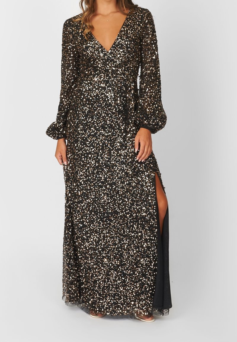 Femme portant une longue robe noire à paillettes avec des manches longues bouffantes et une fente latérale haute jusqu'à la cuisse.