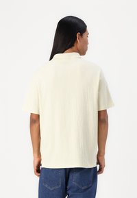 Polo à manches courtes, couleur jaune pâle, avec un tissu texturé, col standard et coupe décontractée, associé à un pantalon en denim bleu.