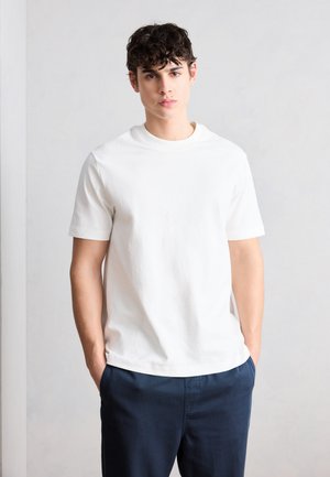 Νεαρός άνδρας με σκούρα σγουρά μαλλιά, φορώντας απλό λευκό κοντομάνικο μπλουζάκι και σκουρό μπλε παντελόνι, στέκεται με τα χέρια στις τσέπες.