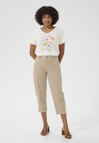 Cremfarbendes V-Ausschnitt-T-Shirt mit floralem Design, kombiniert mit beigen Caprihosen. Braune Absatzsandalen runden den Look ab. Durchgehend glatte Textur.