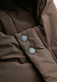 Tessuto imbottito marrone con una superficie liscia, caratterizzato da due bottoni neri rotondi. Il design presenta pieghe e una forma morbida e avvolgente.