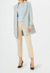 Manteau à motifs pied-de-poule en bleu et blanc, associé à un pull bleu clair et un pantalon beige taillé. Des escarpins noirs complètent la tenue.