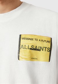 Hvid bomulds t-shirt med en rektangulær gul trykt lap, der har sort tekst: "BESKED TIL EN FREMTID," "ALLSAINTS," og "ADVARSEL."