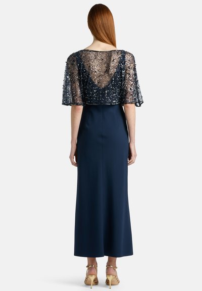 Mornarska obleka s prosojnim prekrivalom s sequins, ki ima dekolte na hrbtu in rokave do sredine. Model nosi zlate sandale z odprtimi prsti.