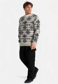 Homme portant un pull beige et bleu marine à motifs, un pantalon noir et des chaussures noires, se tenant contre un fond blanc, regardant sur le côté.