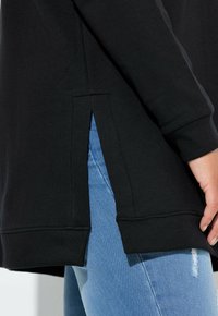 Vista laterale di una persona che indossa una maglietta nera a maniche lunghe con fentature laterali e jeans di denim azzurro chiaro con lievi strappi sulla coscia.