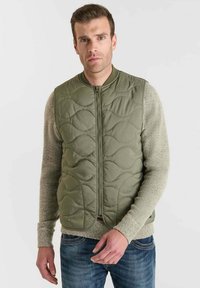 Olijfgroen gewatteerd vest met een voorrits, voorzien van een ronde kraag en een gestructureerde stof. Draagt over een lichtgrijze gebreide trui.