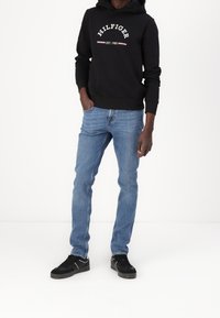 Felpa nera con il logo "HILFIGER EST. 1985", abbinata a jeans blu e sneakers nere con dettagli bianchi. Presentazione di un outfit casual.