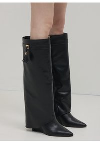 Černá kožená vysoká bota se širokým holením, špičatou špičkou a zlatými kovovými detaily. Disponuje bočním zipem a ozdobným střapcem.