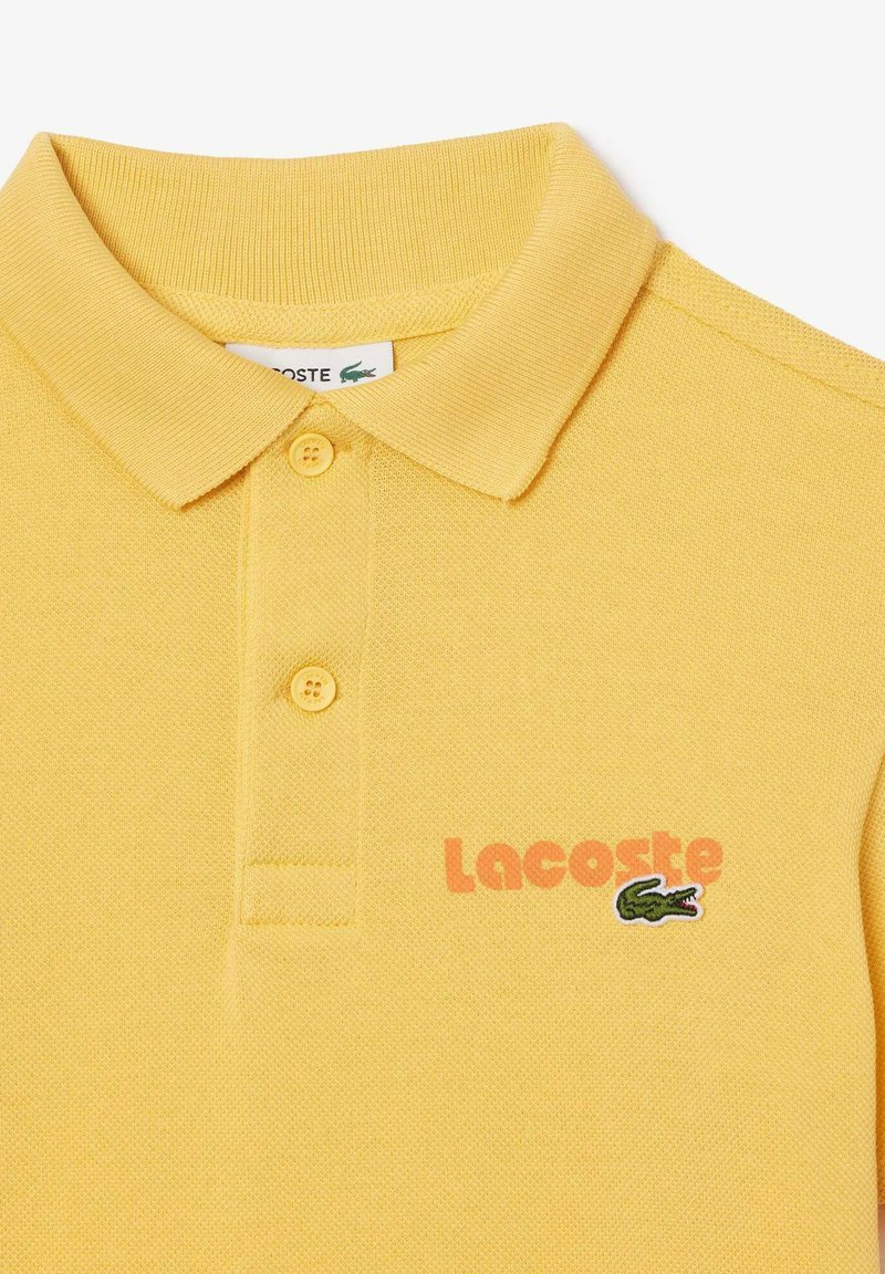 Polo shirt jaune avec un motif texturé, patte de boutonnage à trois boutons, logo crocodile vert brodé et texte "Lacoste" orange.