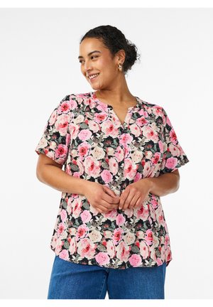 Kvinde smilende og kiggende sidelæns iført en kortærmet blomstret bluse med pink og røde roser samt blå jeans, hænderne foldet foran.
