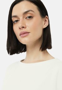 Weißes Oberteil mit weitem Ausschnitt, glattem Stoff und kurzen Ärmeln. Merkmale sind schulterlanges dunkles Haar, dezentes Make-up und eine nahe Gesichtsperspektive.