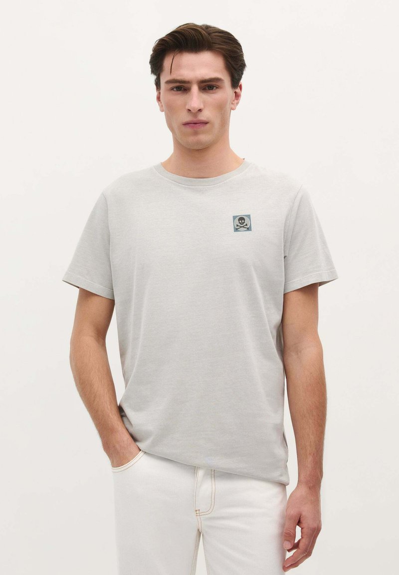 Grijze katoenen t-shirt met korte mouwen, voorzien van een kleine blauwe vierkante patch met een doodskop en gekruiste botten ontwerp op de borst.