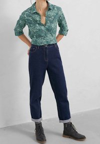 Camicia a maniche lunghe verde a fantasia, jeans blu scuro a vita alta con orli arrotolati e stivali alla caviglia neri con lacci. Tessuto morbido, vestibilità casual.