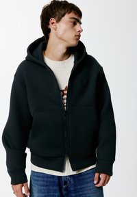 Felpa nera con zip realizzata in tessuto morbido, dotata di un ampio cappuccio, tasche frontali e polsini a coste. Sotto, un maglione crema con un motivo testurizzato.
