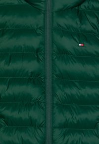 Mørkegrøn puffjakke med lynlås foran. Har horisontale syninger, en glat tekstur og et lille emblem i rød, hvid og blå.