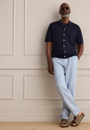 Mann mit Sonnenbrille, kurzärmeligem dunkelblauem Hemd mit Knöpfen, hellblauer Hose und braunen Sandalen, der an einer beige getäfelten Wand lehnt.