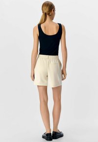 Zwarte tanktop en beige shorts. De shorts hebben een elastische tailleband en zijzakken. Gladde stof met een ontspannen pasvorm. Zwarte schoenen.