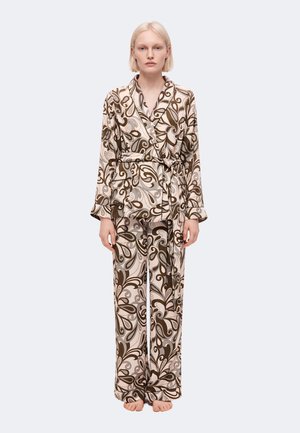 Femme debout, pieds nus, portant un pantalon ample assorti et une veste ceinturée avec un motif abstrait tourbillonnant marron et beige sur fond clair.