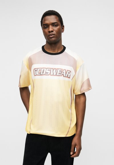 Άνδρας με κίτρινο και μπεζ κοντομάνικο πουκάμισο με το κείμενο «GODSWEAR», στέκεται με το ένα χέρι στην τσέπη μπροστά σε απλό γκρι φόντο.