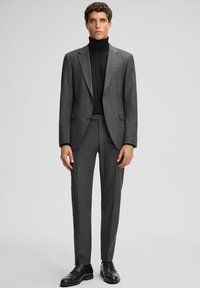 Mann steht in einem grauen Nadelstreifenanzug mit einem schwarzen Rollkragenpullover und schwarzen Lederdressschuhen vor einem schlichten grauen Hintergrund.