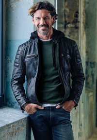 Mann mit lockigem Haar und Bart, der eine schwarze Lederjacke und einen grünen Pullover trägt und sich in einem rustikalen Raum an eine Fensterbank lehnt.