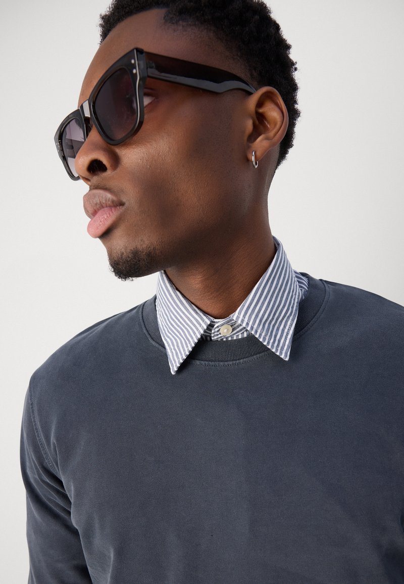 Schwarze Sonnenbrille, grauer Sweatshirt mit Rundhalsausschnitt und ein blau gestreiftes Hemd mit Button-down-Kragen. Glatte Texturen und kontrastierende Farben.