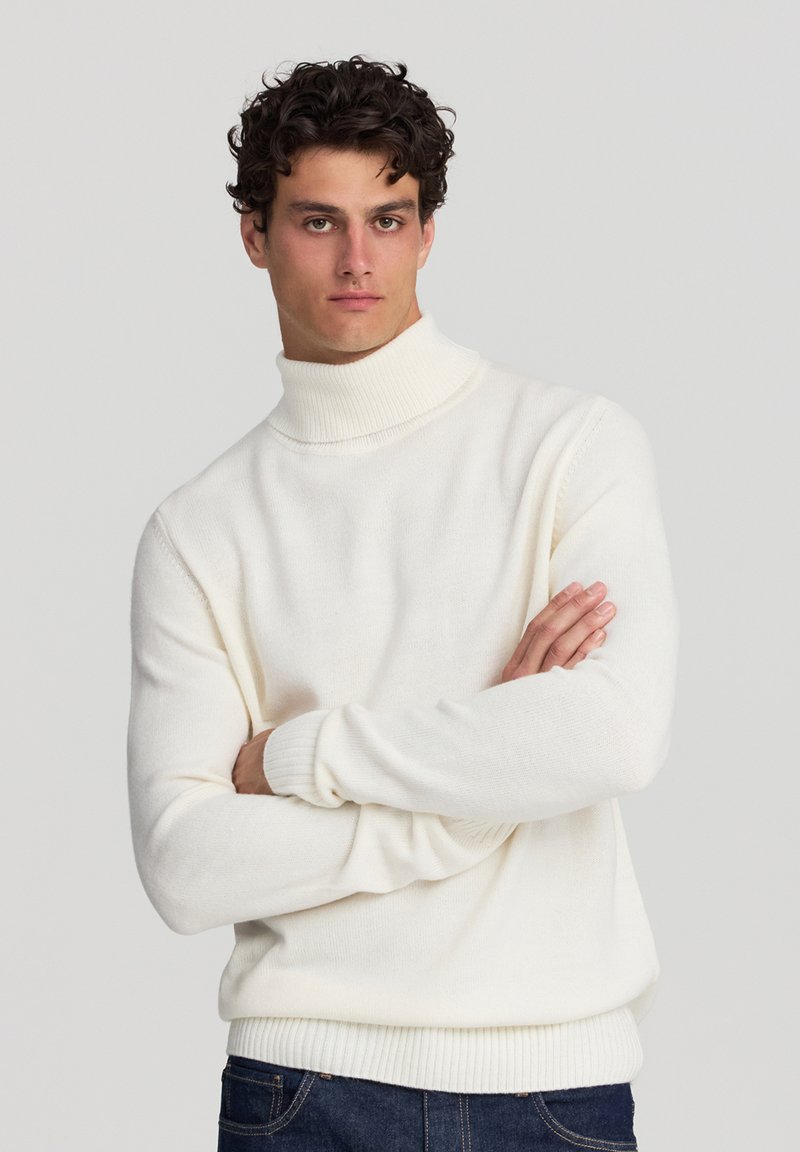 Maglione a collo alto bianco realizzato in materiale morbido e testurizzato. Presenta maniche lunghe e polsini e orlo a coste, abbinato a jeans di denim scuro.