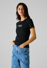 Schwarzes, geripptes kurzärmliges T-Shirt mit dem in Weiß gestickten Logo "RED BULL RACING". Getragen mit ausgebleichten blauen High-Waist-Jeans.