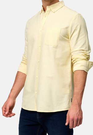 Homme portant une chemise boutonnée à manches longues jaune pâle avec une poche poitrine et un jean bleu foncé sur un fond uni.