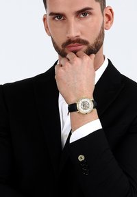 Analoge polshorloge met een goudkleurige kast, transparante wijzerplaat, zwarte leren band en zilveren accenten. Draagt op een hand tegen een donker pak.